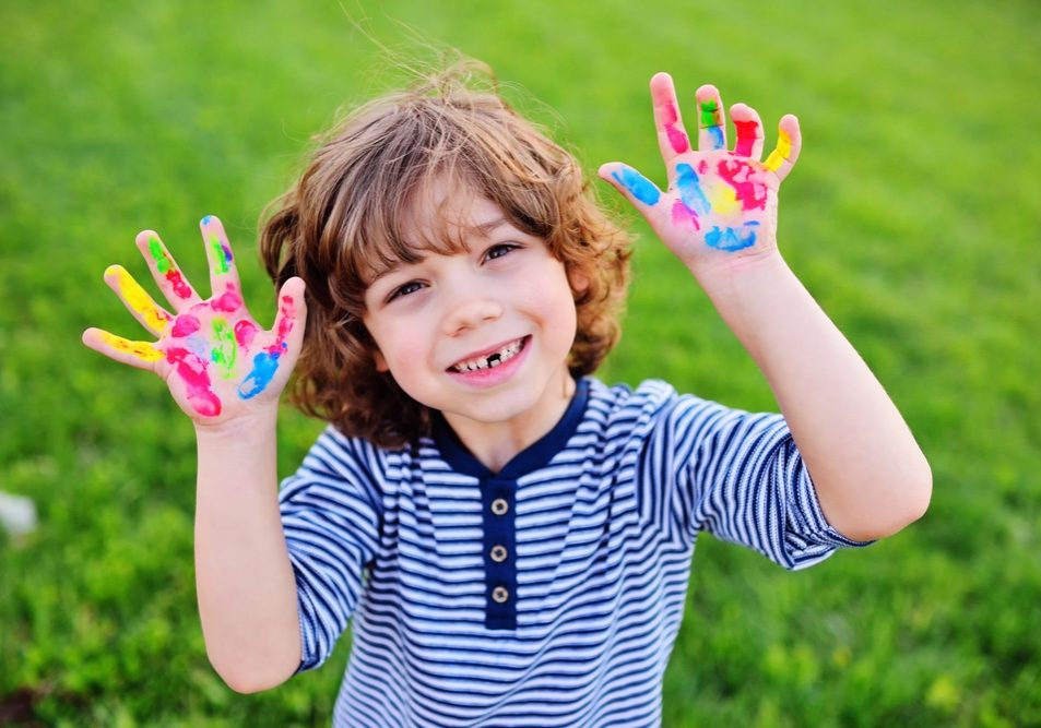 cheerful child boy with curly hair without front milk tooth shows hands dirty with multi-colored finger paints and smiles.